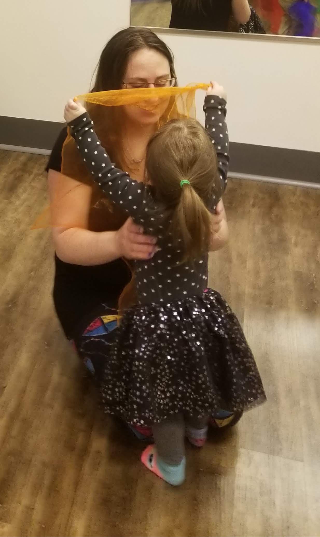 A mom kneeling down playing peek-a-boo with a golden scarf with her little girl during Zumbini